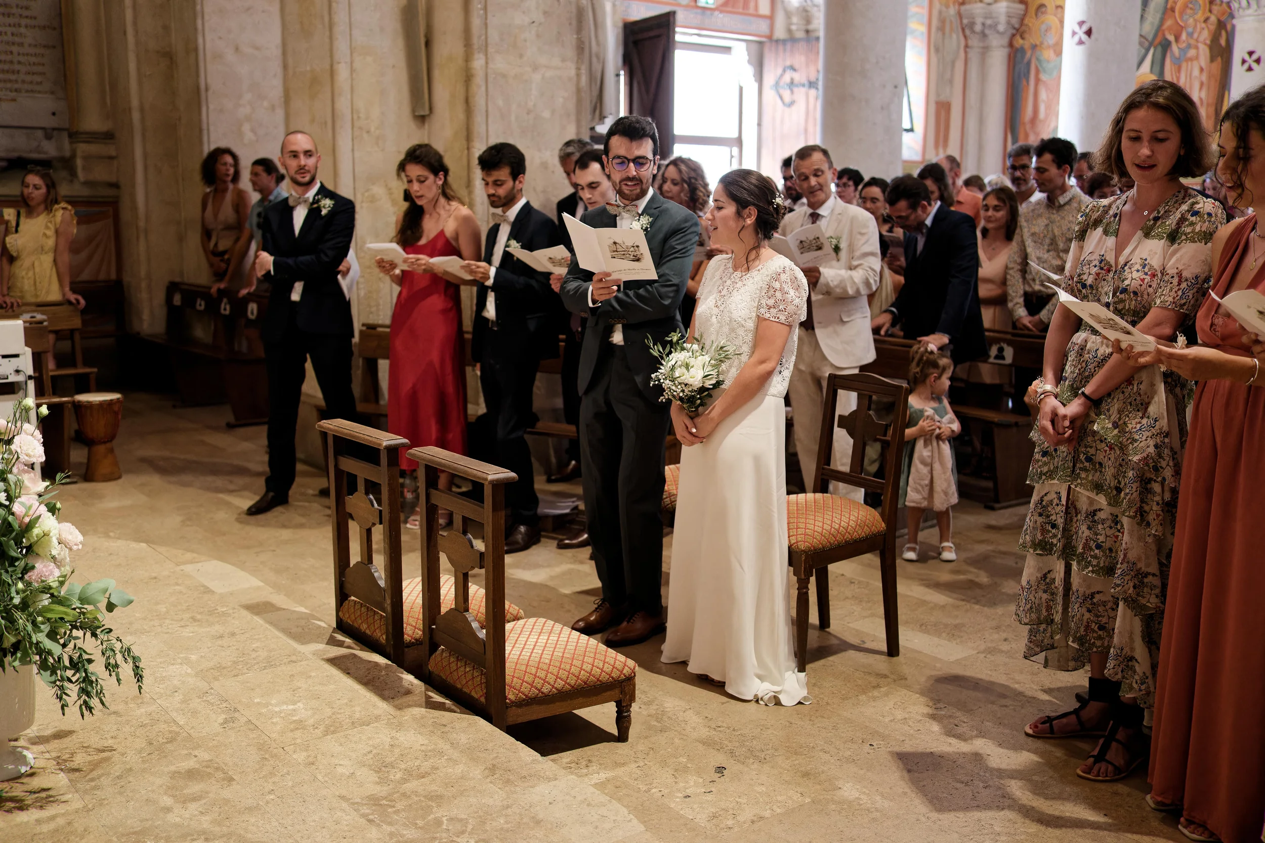 Les mariés et leurs invités suivent les chants dans leurs livrets, près des colonnes et des fresques de l’église.