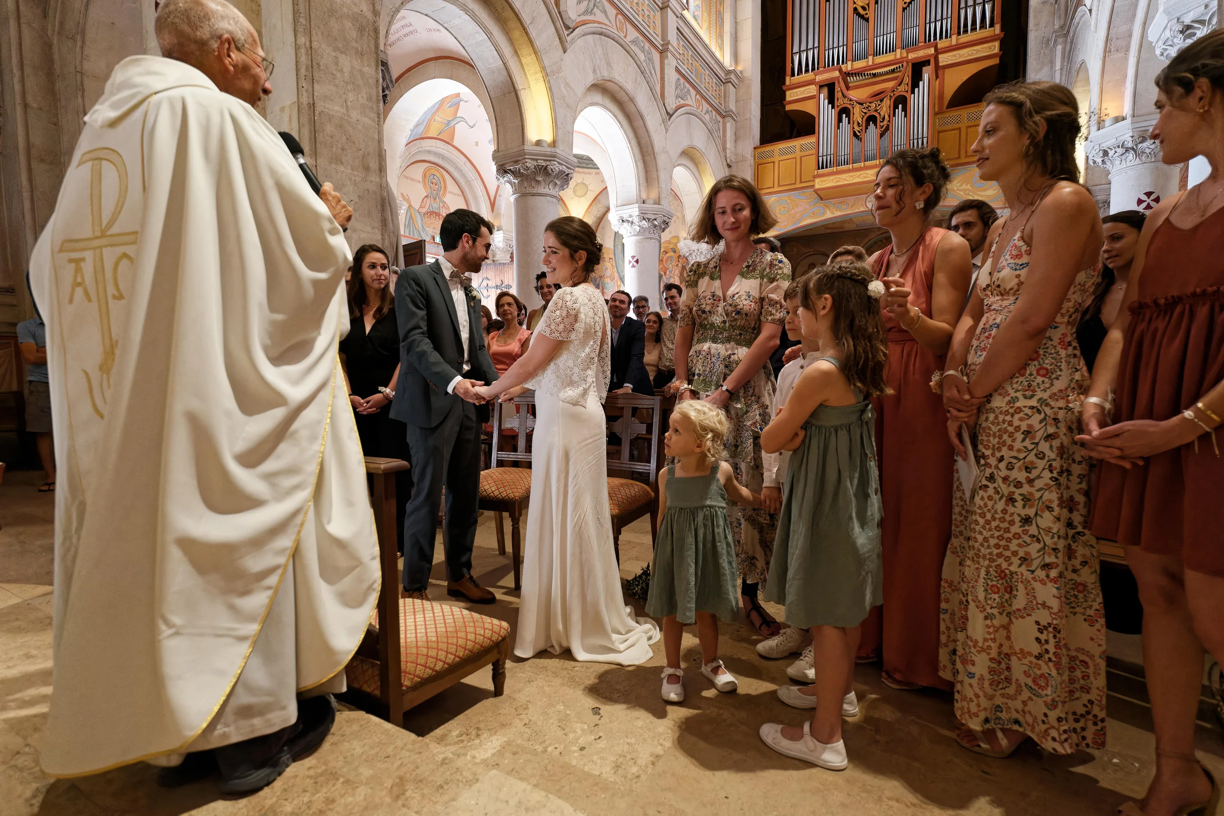 Les mariés se tiennent les mains face au prêtre, entourés de leurs proches et de deux enfants, sous les arches de l’église.